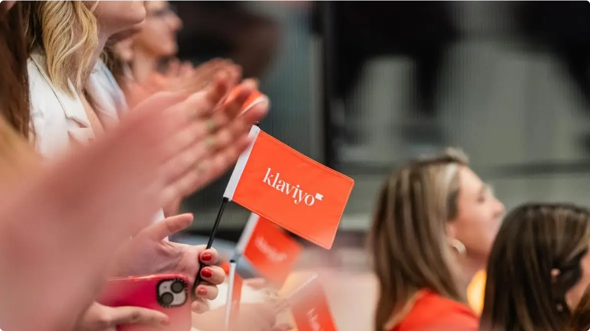 Focus on a small handheld Klaviyo flag. Out-of-focus background shows several Klaviyo employees, each holding such a flag.